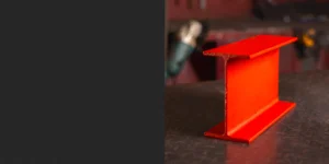 Red universal beam resting on a sleek black counter, showcasing its strong and versatile design. The bold red finish highlights its durability and suitability for structural applications. This universal beam is ideal for projects requiring strength and reliability in construction.