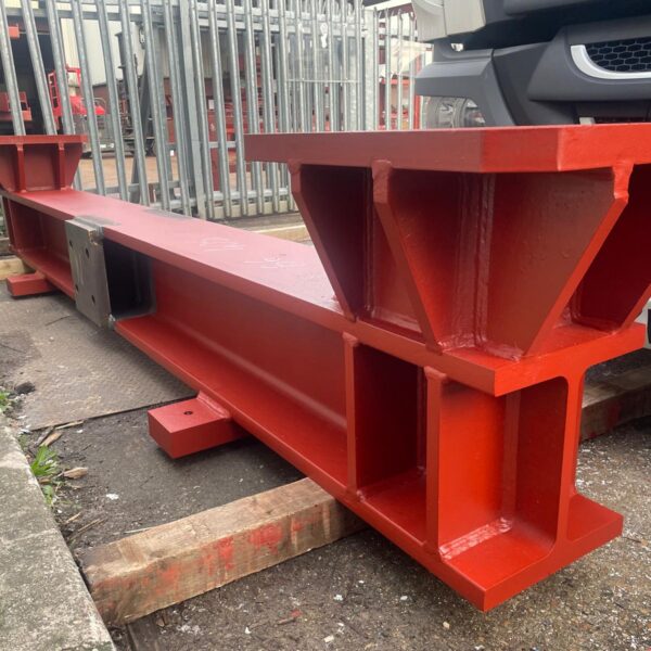Red oxide–primed structural steel column with heavy-duty baseplate and pre-drilled connection holes, staged for transport at an industrial site.